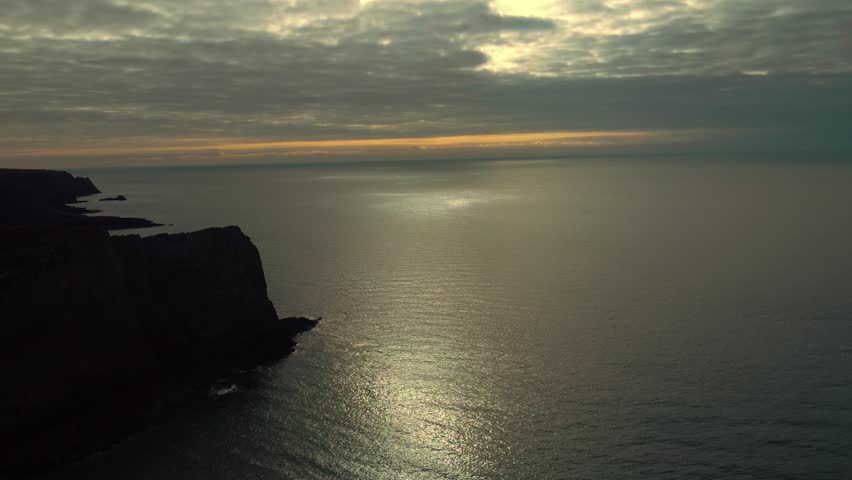 Panning Aerial View of Welsh Coastline with Bright Sun Reflecting on Ocean Behind Dark Clouds with Dynamic Rugged Coastline.