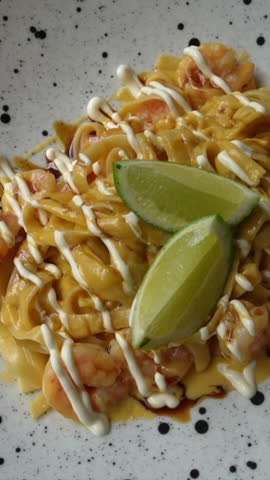 Vertical video capturing a close-up shot of flavorful Colombian shrimp pasta garnished with lime and drizzled sauce. Bright lighting enhances the delectable presentation.