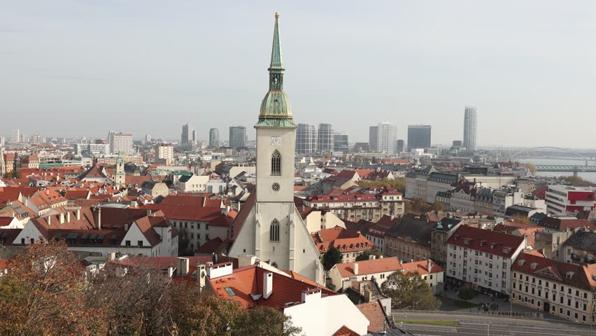 Scenic autumn view of Bratislava with medieval St. Martin Cathedral rising above historic terracotta tiled rooftops against backdrop of modern skyscrapers on bank of Danube River, Slovakia