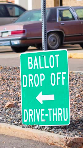 A clear election sign reads “Ballot Drop Off” with an arrow, standing near a road, symbolizing safe, secure, and accessible civic duty, democracy, equality, and freedom to participate. Vertical Video