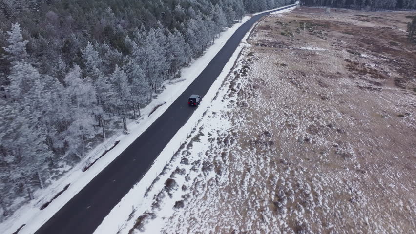 Aerial view of black car driving along mountain pass. Vehicle travels on a street that separates a snowy pine forest from a bare field