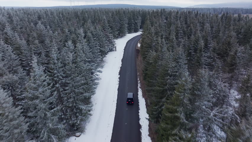 Aerial footage of car driving on a curvy road through a dense, snow covered pine forest. Wintry landscape reveals mountain scenery