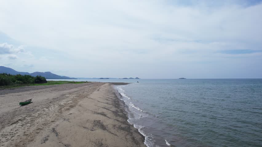 Aerial drone footage flying over a long, empty beach. A small green traditional fishing boat (jukung) is on the sand. The ocean is calm. Indonesia.