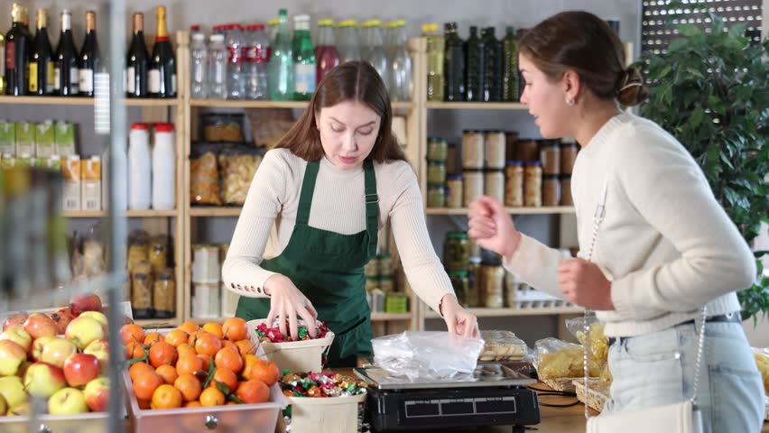 Grocery store selling area. Female employee sells candies to woman, puts them in bag, puts them on scale and weighs sweets. Brunette European girl buys candies by weight.