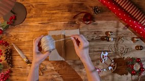 Top view of a woman hands cutting twine with gold scissors while wrapping a Christmas gift in kraft paper. A cozy holiday scene with festive decorations, a pine cone, ribbon, and wrapping paper. - Powered by Shutterstock - Get 15% off with code: PIKWIZARD15