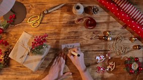 Top view of hands writing Merry Christmas on a kraft paper gift tag next to a wrapped gift, ribbons, and decorations on a wooden table. Cozy holiday packaging and a handmade greeting concept. - Powered by Shutterstock - Get 15% off with code: PIKWIZARD15