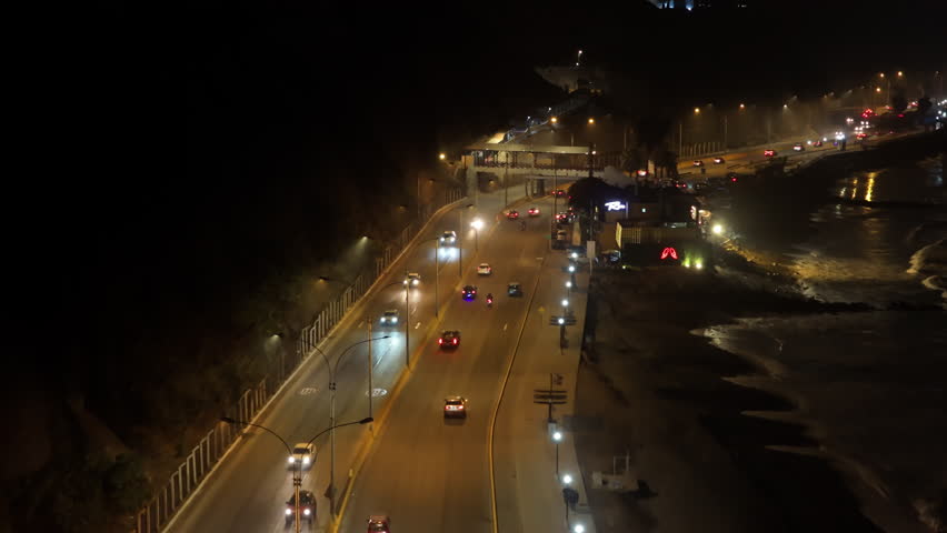 Nighttime aerial shot following cars driving on Circuito de Playas highway along coast in Lima, Peru, with city lights illuminating the road