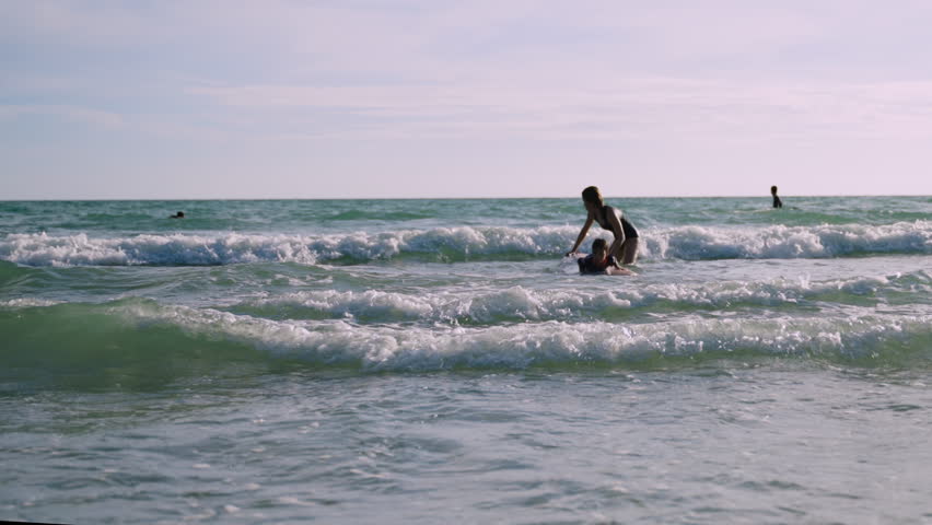 Happy little boy learning to play with a surfboard from his mother, having fun and passion in the sea during summer vacation at the beach. An Asian mom teaches and trains her cute son on how to surf. 
