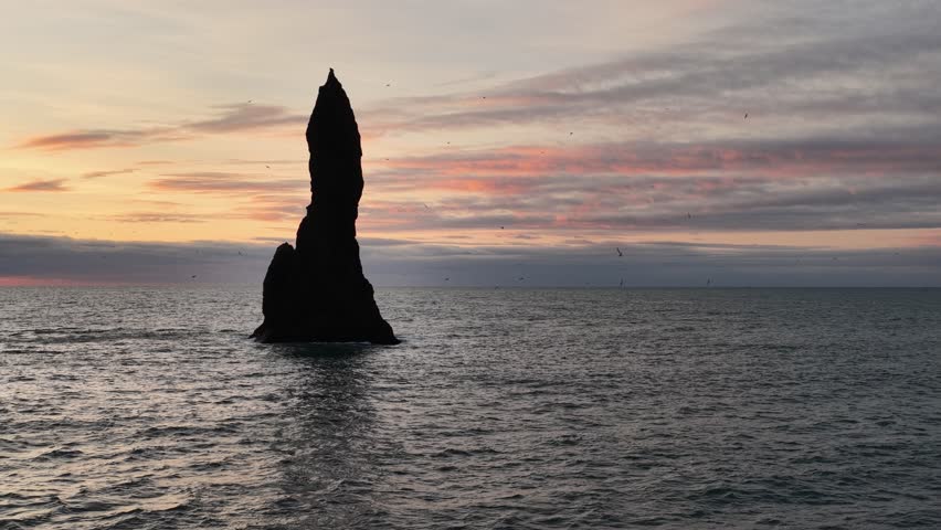 Aerial view of a dramatic dark rock formation rising from the sea, set against a backdrop of a colorful sky at dusk, Iceland.