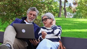 Happy senior couple sitting on a park bench and using a laptop computer. The mature man and woman are smiling, talking, and laughing together - Powered by Shutterstock - Get 15% off with code: PIKWIZARD15