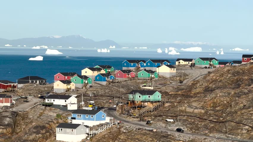 Brightly colored houses stand near the ocean, surrounded by icebergs. The cloudy sky adds a unique ambiance to this tranquil Greenlandic landscape. Nature and culture blend beautifully here.