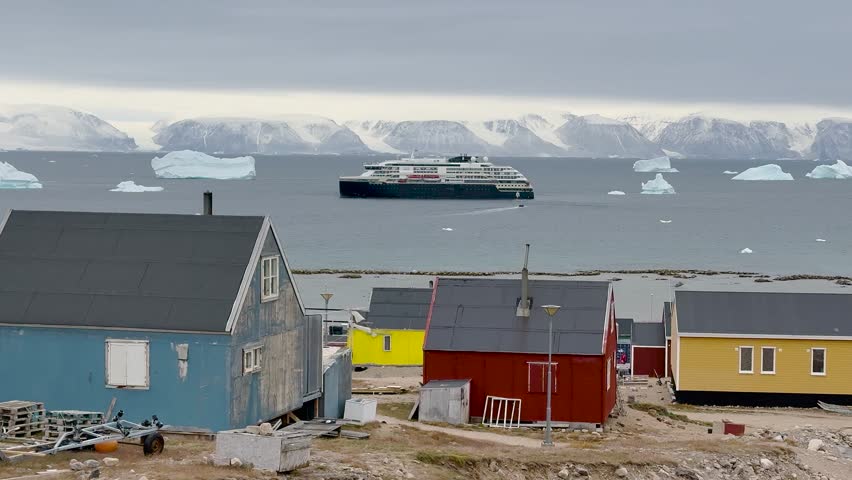 Brightly colored houses stand near the ocean, surrounded by icebergs. The cloudy sky adds a unique ambiance to this tranquil Greenlandic landscape. Nature and culture blend beautifully here.