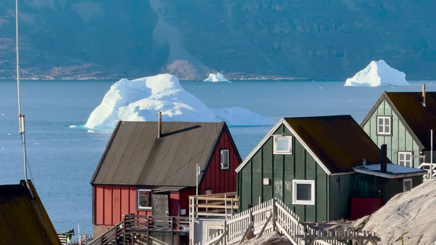 Brightly colored houses stand near the ocean, surrounded by icebergs. The cloudy sky adds a unique ambiance to this tranquil Greenlandic landscape. Nature and culture blend beautifully here.