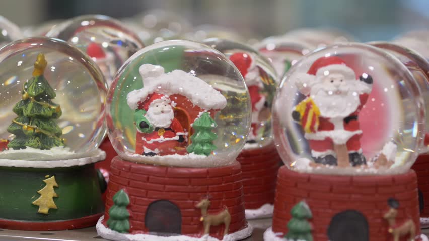 Assorted snow globes with trees and santas closeup of colorful shelf display, vendor arranging variety of holiday collectibles, sparkling snowfall inside glass, joyful seasonal market scene