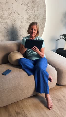 A woman joyfully interacts with a tablet while seated on a modern sofa, showcasing comfort and technology.
