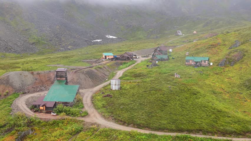 Aerial view of buildings on grassy hillsides with a dirt road winding through, the scene is shrouded in mist and fog, Palmer, Alaska, United States.