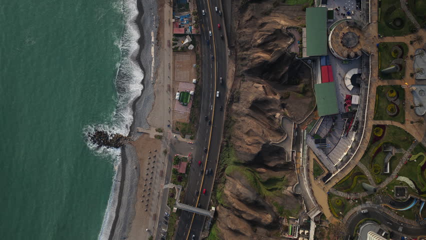 Aerial top down perspective of Lima, Peru, showing the Pacific Ocean waves, a pebble beach, cliffside road, and urban park