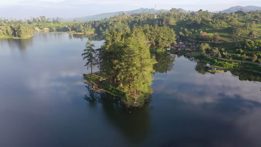 Cinematic footage of a lake at sunrise with golden morning light reflecting on the surface. Surrounded by green hills and trees, the serene atmosphere captures the calm beginning of the day in a tropi