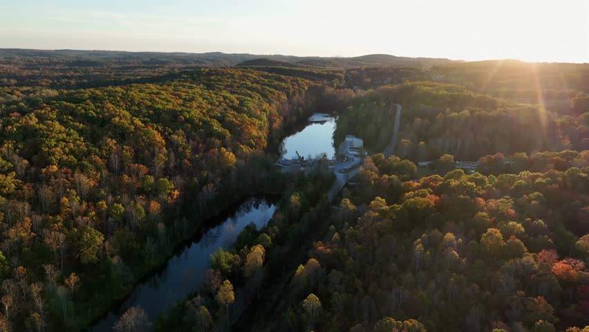 Aerial wide shot of tranquil river between dense colored forest trees during golden sunset. Peaceful atmosphere in fall season. Virginia, USA.