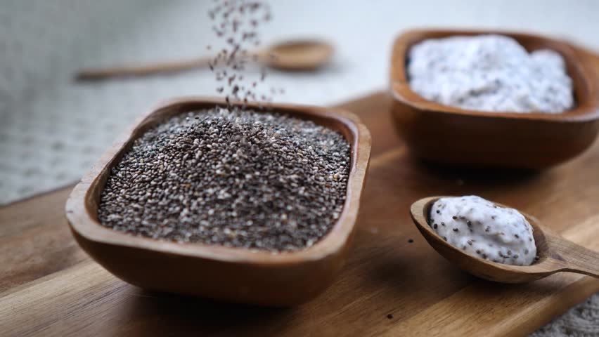 Slow motion close up of healthy chia seeds pouring into a rustic wooden bowl, with prepared creamy pudding on a spoon nearby, showcasing a nutritious superfood breakfast ingredient