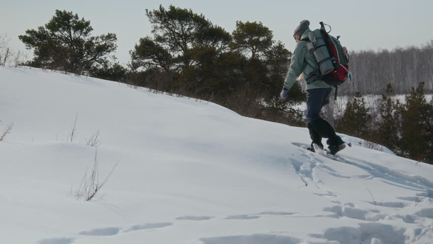 Wide angle side view shot of young woman hiking alone in winter and walking uphill in snowshoes carrying big backpack, copy space