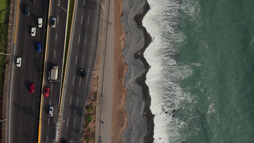 Top down aerial view of vehicles driving along a scenic coastal highway next to the Pacific Ocean with waves crashing on a pebble beach in Lima, Peru
