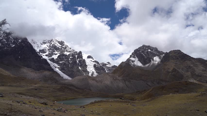 A wide, stationary shot of the sacred, snow-capped Nevado Ausangate mountain in Peru. One of the Seven Glacial Lakes sits below its peak under a blue sky with scattered clouds