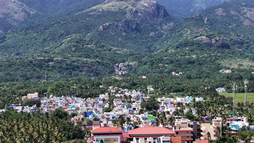 Aerial view of a village nestled at the foothills of a majestic mountain range, the Western Ghats in South India. The town create a striking contrast against the deep green forests, lush paddy fields