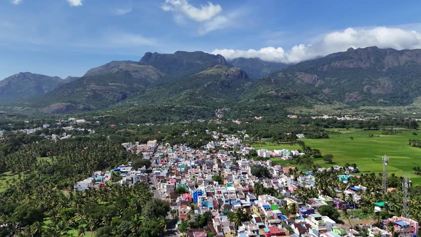 Aerial view of a vibrant South Indian town nestled directly at the foothills of a majestic mountain range. The foreground features a dense cluster of colorful houses and a vivid green paddy field