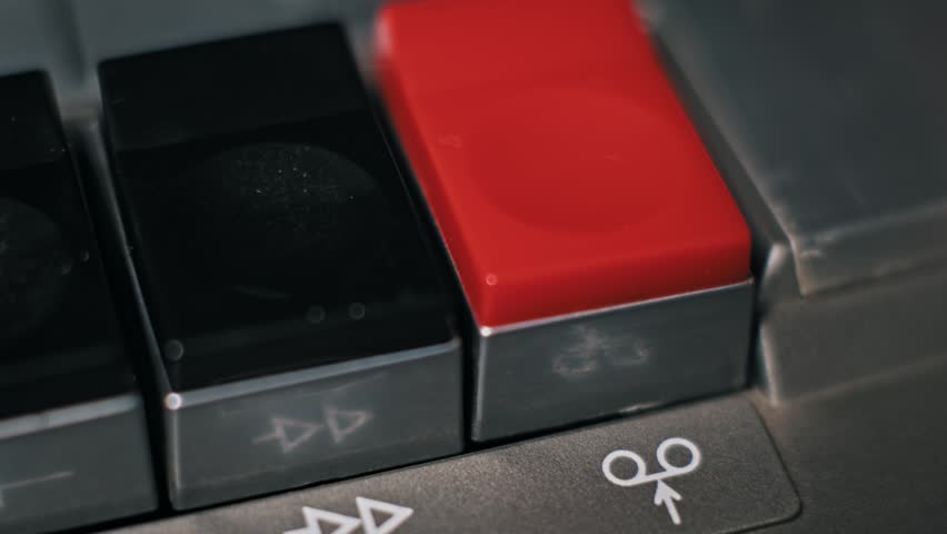 Extreme close-up view of a finger depressing the red record key on a retro audio cassette player. The button is pushed down into the metal control panel to begin capturing sound on analog tape.