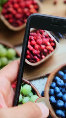 Influencer hand holding a modern smartphone and recording a video or taking photos of a healthy flat lay composition with fresh organic berries for social media stories or a food blog post
