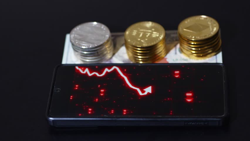 A smartphone show a red bearish trading chart beside a stack of Bitcoin coins and USD 100 bills. Financial loss, risk, and the volatile relationship between cryptocurrency and traditional fiat money.