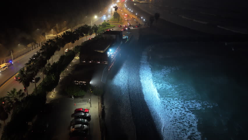 Aerial view of illuminated coastline in Lima, Peru, at night. Cars driving along Circuito de Playas road with ocean waves crashing