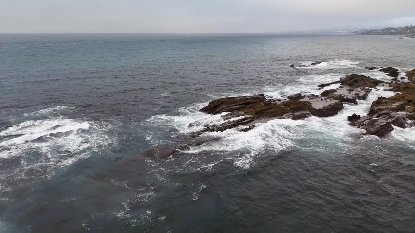 Drone view of gentle waves breaking over rugged rocks under a hazy coastal sky