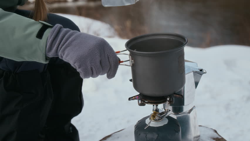 Close up shot of unrecognizable young woman as female hiker putting meal pouch into boiling pot using portable gas stove while taking a break at camping stop in winter