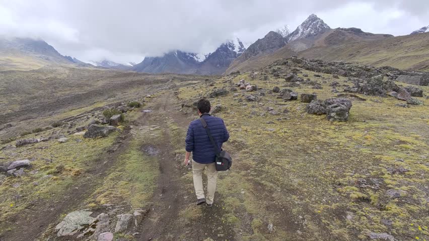 From behind as TV host Charles Huang walks a dirt path toward snow-capped mountains on his trek to the 7 Lakes of Ausangate