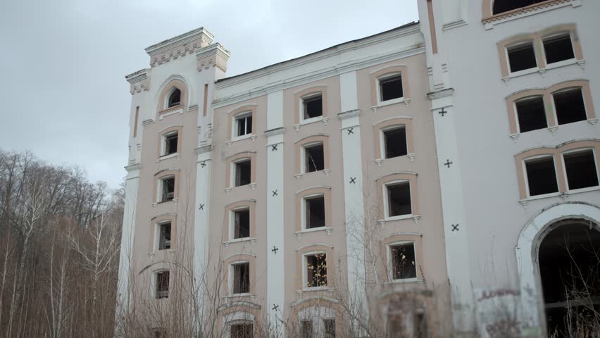 Abandoned hospital or hotel, pale facade with broken windows, ghostly figure stands motionless in single window, cold sky and leafless trees add mystery and sense of urban legend.