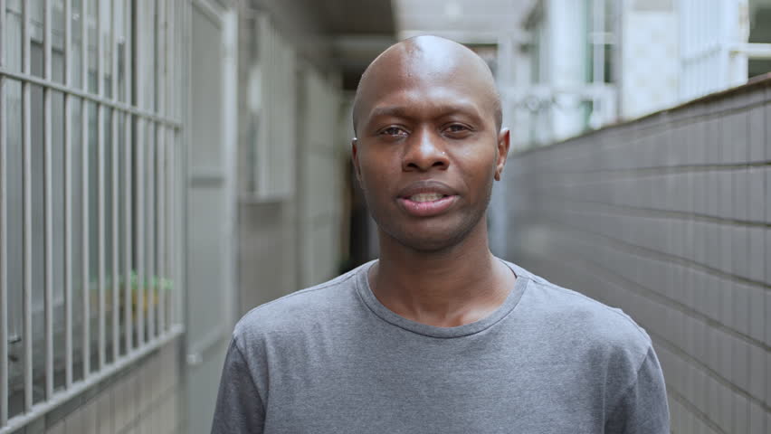 Man In Gray Shirt Standing In Narrow Hallway With Tiled Wall Looking At Camera With Neutral Expression, Centered Frame In Outdoor Location