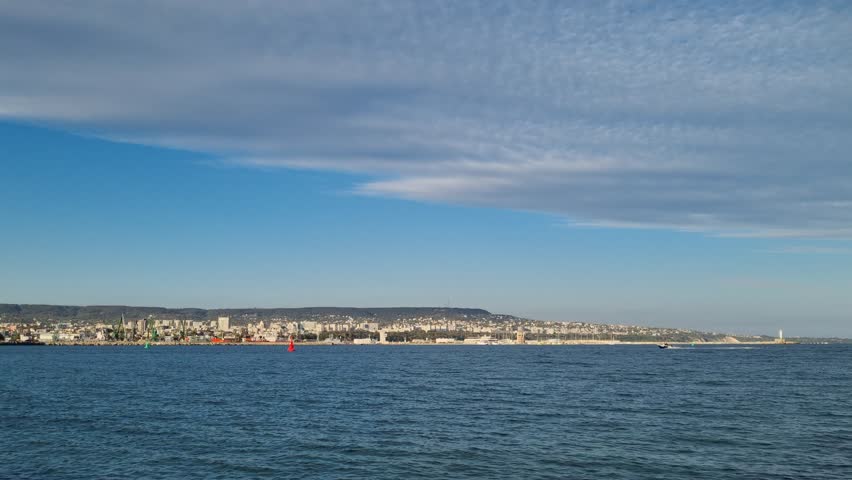 City coastline view across calm blue sea under clear sky. Peaceful urban seaside landscape with distant buildings, horizon, and sunlight reflection. Ideal for travel, tourism, and cinematic footage.