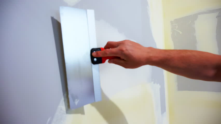 Construction worker spreading joint compound on drywall wall corner using a metal taping knife. Wall smoothed and finished as part of a home renovation.