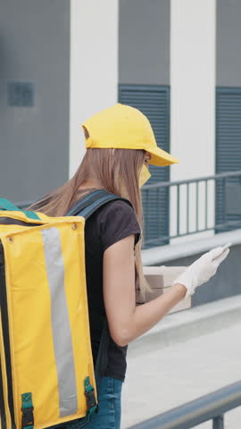 A woman wearing a yellow cap and mask is delivering food, holding packages, and wearing gloves. She has a yellow delivery bag.