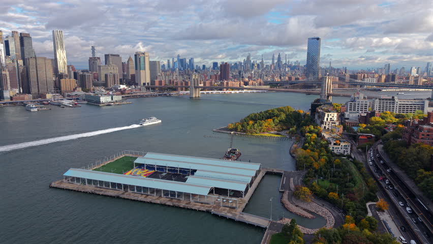 Stunning aerial shot of Manhattan New York skyline across the river, with ferry in motion, Brooklyn bridge waterfront park, and pier structure under a dynamic cloudy sky in New York City.