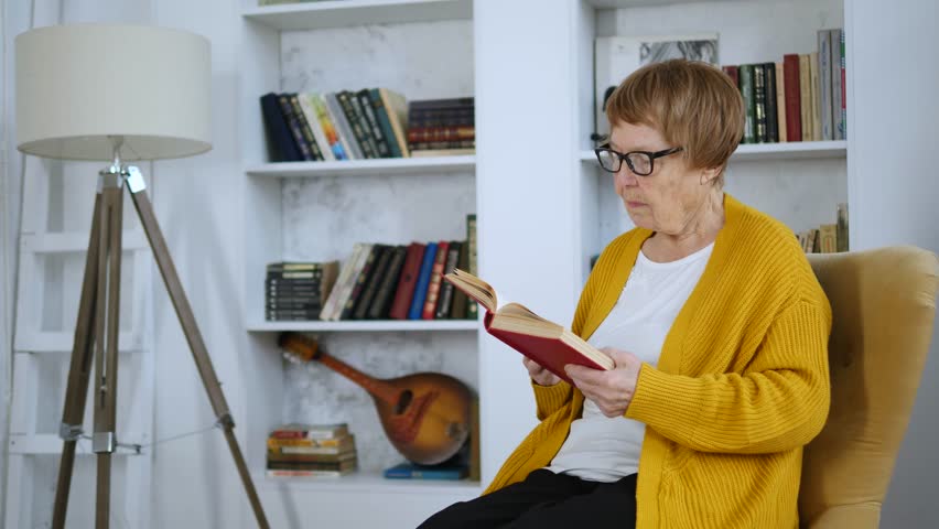 Senior woman with glasses relaxing in a comfortable armchair while reading a novel in her cozy living room. The calm and peaceful atmosphere of a home library promotes education and mental well being