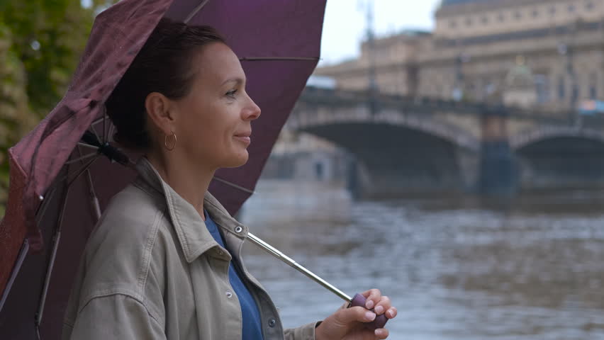 Woman in the rain in Prague. Woman standing with colorful umbrella overlooking river, elegant cityscape of prague featuring iconic bridge and architectural buildings during cloudy autumn day