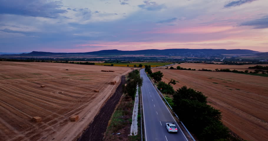 Sunset Aerial Landscape Panoramic view of  Fast Modern White Car Driving On a Rural Highway Route Through an Agricultural Fields
