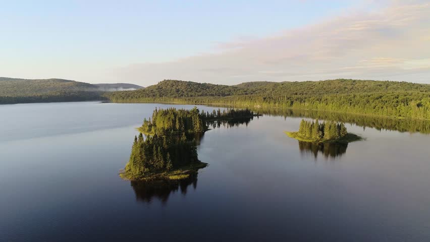Dynamic Aerial Skimming Island Treetops. A dynamic drone shot flies fast and low over the water, passing just above the treetops of small islands in a pristine lake at golden hour.