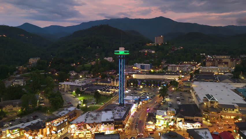 Aerial panorama of Gatlinburg, Tennessee at sundown with bustling main street filled with shops and attractions, observation tower and Smoky Mountains National Park nearby.