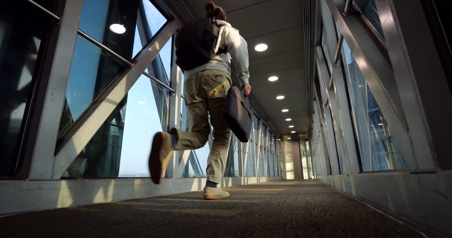 Low-angle wide slow-motion shot of male traveler with backpack and laptop bag hurrying and running along empty airport jet bridge toward airplane. Final call and last-minute boarding rush scene