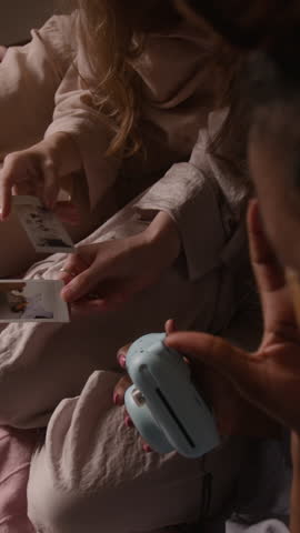 Vertical close-up of three diverse young girls with cosmetic eye patches sitting close together on bed, watching instant photos developing, during fun pajama party