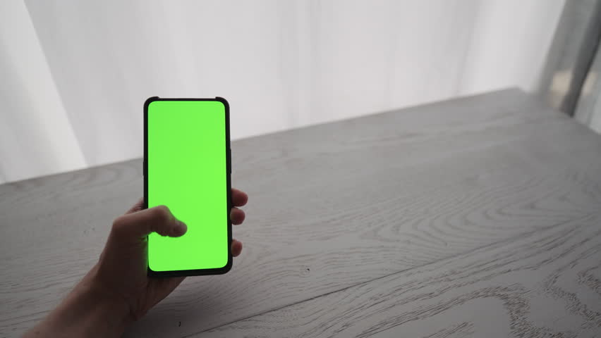 Man showing and using smarphone with green screen while sitting behind white oak table